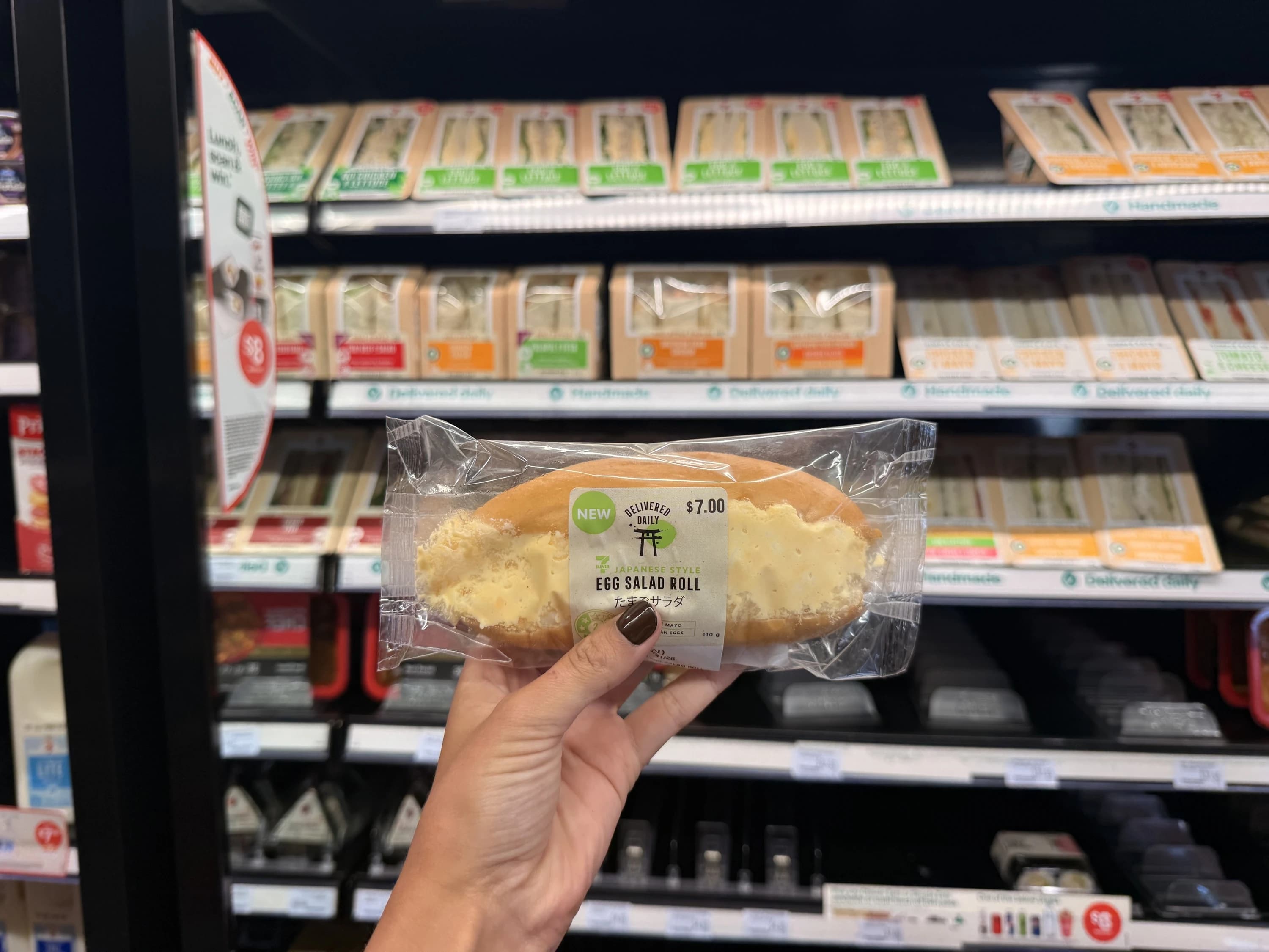 豪州セブンの卵サラダロール。ロールパンに卵サラダを挟んだ商品を、サンドイッチ売り場の前で手に持っている写真